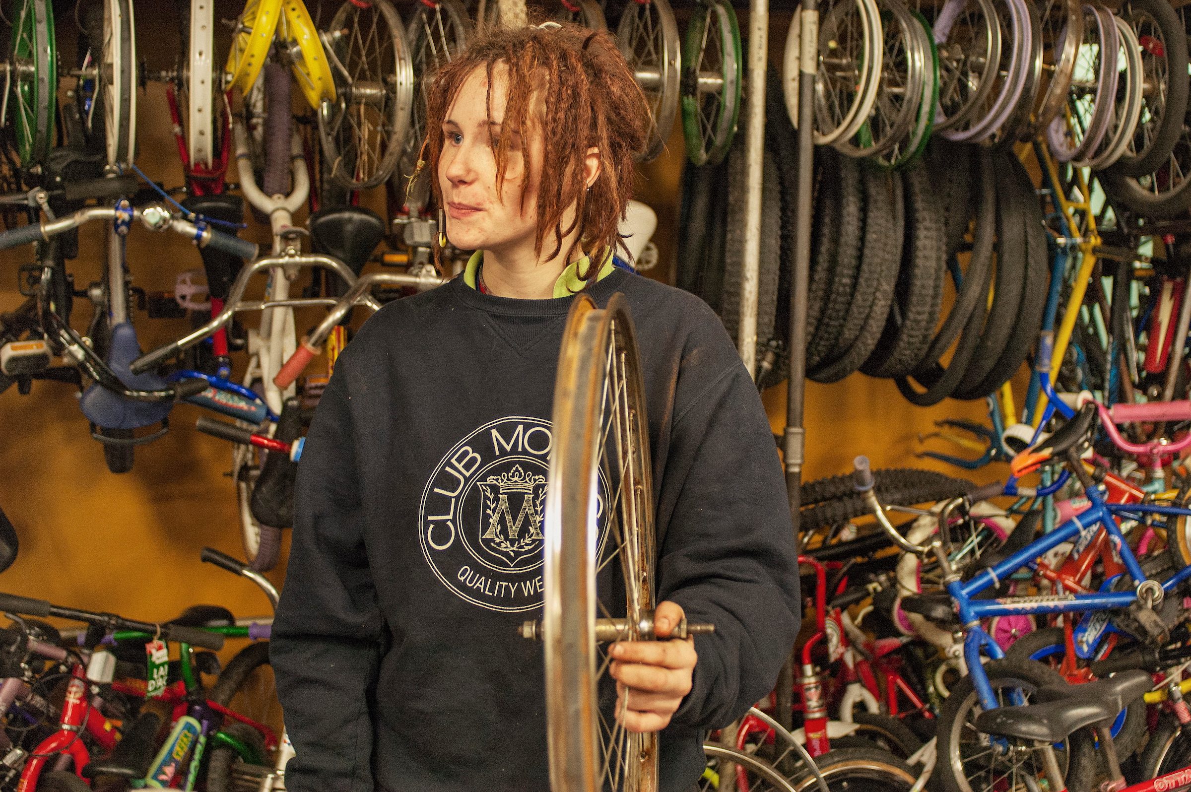 A volunteer holding a bicycle wheel in the Recycle Cycles Community Bike Shop, surrounded by a variety of bike wheels and frames.