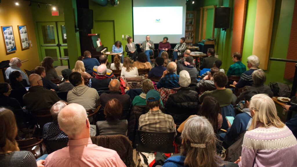 A diverse audience attending a panel discussion at the Commons, with panelists seated in front of a screen. Caption: The Commons hosts an engaging panel discussion, bringing community members together to explore ideas, share knowledge, and inspire collective action.