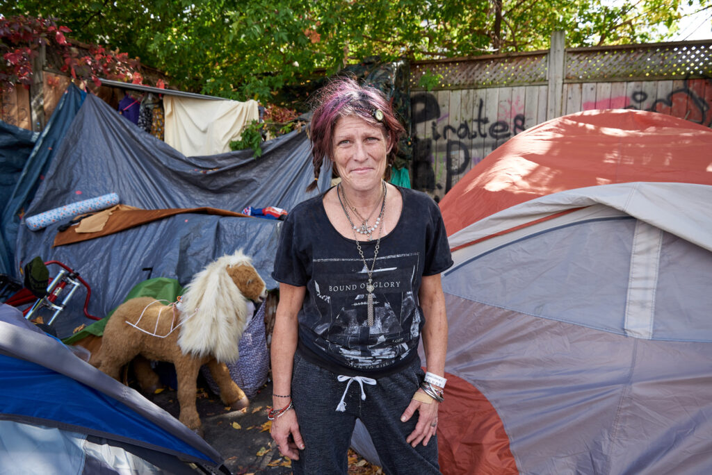 A woman stands in front of her tent in a vibrant encampment, surrounded by personal belongings and makeshift structures in Kitchener, Ontario.