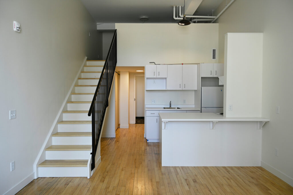 Interior of Unit 204 at Queen Street Apartments, featuring a staircase, kitchen area, and bright wooden flooring. A supportive housing project by The Working Centre, Kitchener.