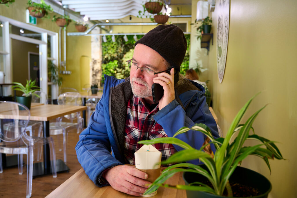 Don Toomey, a Working Centre staff member, enjoying a coffee while on a call at Fresh Ground Cafe, surrounded by greenery and a cozy ambiance in Kitchener, Ontario.