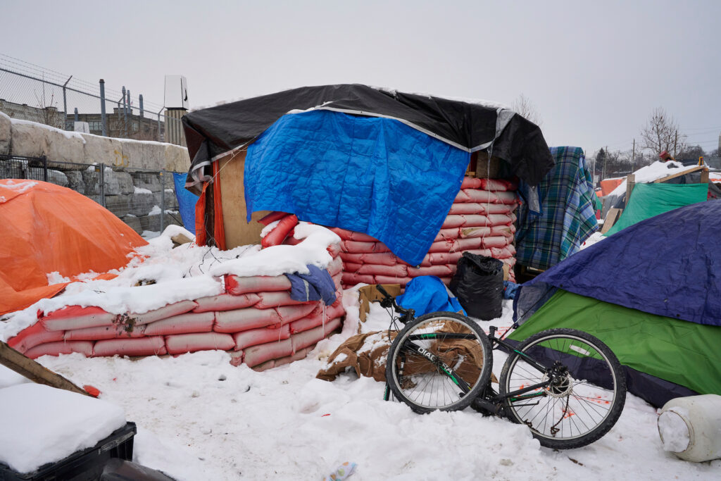 A snowy winter view of the Victoria Weber tent encampment in Kitchener, Ontario, featuring makeshift shelters reinforced with sandbags, tarps, and a bicycle in the foreground.