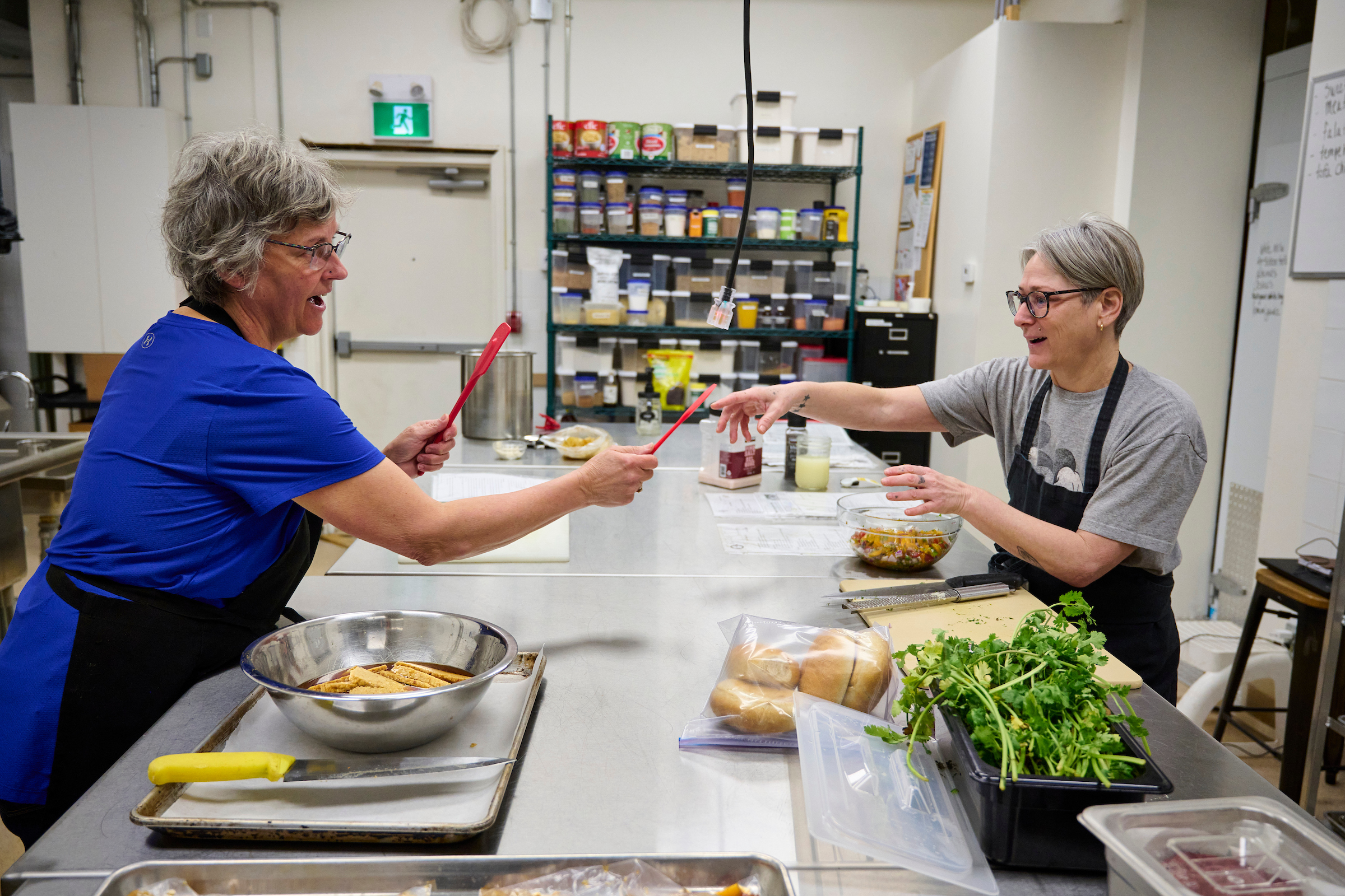 Erin and Jane, staff members of Fresh Ground Café in Kitchener, Ontario, working together in the kitchen preparing plant-based meals.