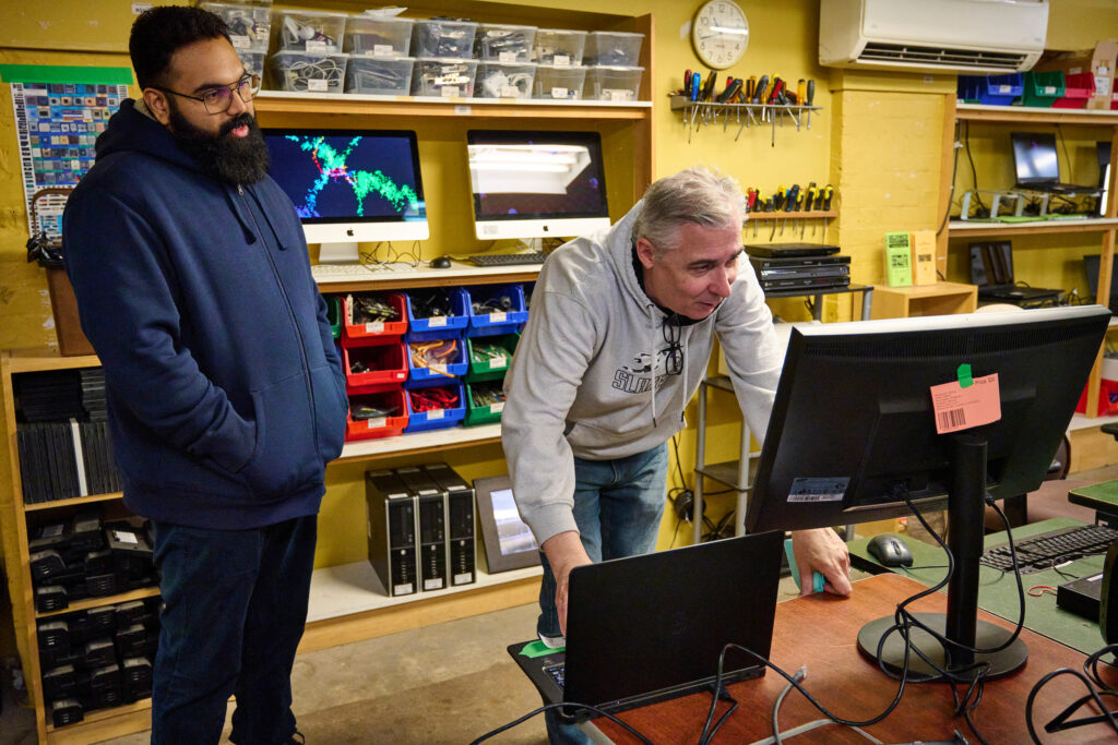 A customer observes Charles, a staff member, testing a computer monitor at the Computer Recycling facility in Kitchener, Ontario.