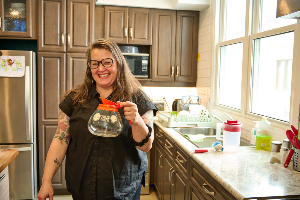 Tanya Roper smiling and holding a coffee pot in the kitchen common room at Hospitality House, Kitchener, Ontario.