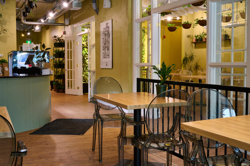 The front entrance area of the newly reopened Fresh Ground Café in Kitchener, Ontario, featuring wooden tables, transparent chairs, green walls, and lush indoor plants.