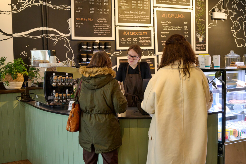 Eva, a staff member at Fresh Ground Café, serves two customers at the newly reopened café in Kitchener, Ontario, with a vibrant black-and-white menu wall in the background.