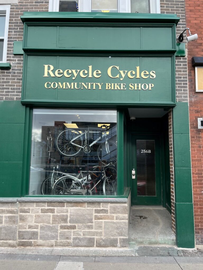 The storefront of Recycle Cycles Community Bike Shop, featuring a green facade with gold lettering and a display of bicycles in the window.