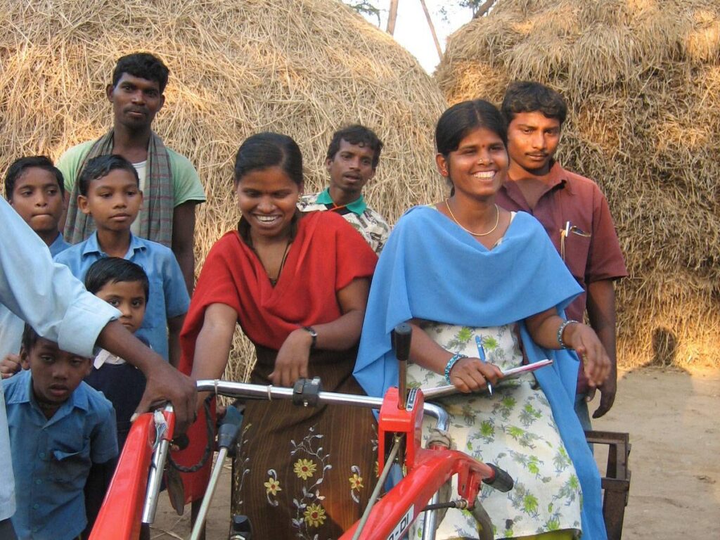 A group of smiling villagers, including women and children, participate in a demonstration of a red power tiller in rural Odisha, India.